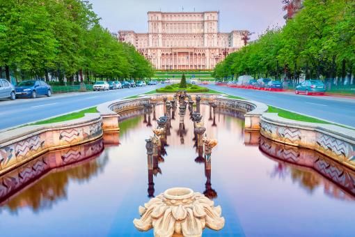 Photograph of fountains along the tree-lined Bulevardul Unirii, a major thoroughfare in central Bucharest, with the Palace of the Parliament in the background at dawn.