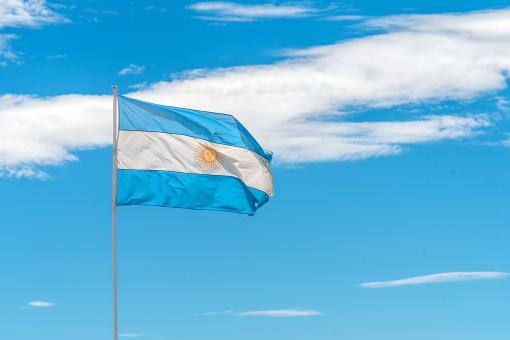 A blue and white argentinian flag with a yellow sun sits on a pole. The sky is clear and blue, with a few clouds scattered throughout. The flag is waving in the breeze, giving the impression of a sunny day.